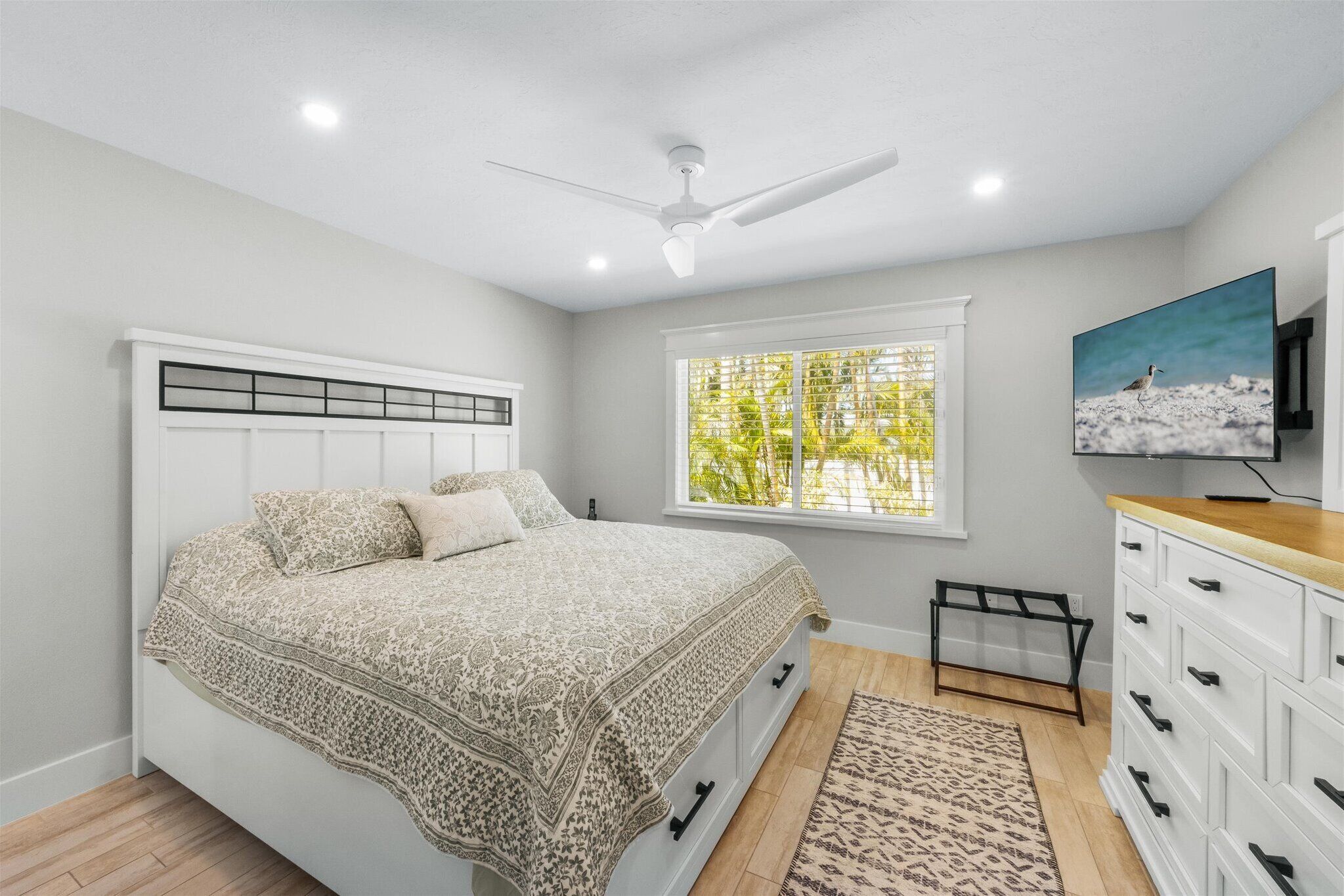 A bedroom with a bed, a ceiling fan, a window with a view of trees, a TV, and a dresser.