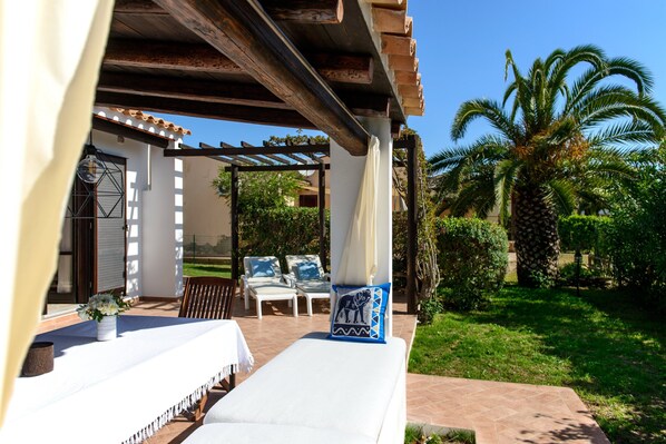 An outdoor seating area with a covered patio, a table with a vase of flowers, and a lounge chair with a decorative pillow.