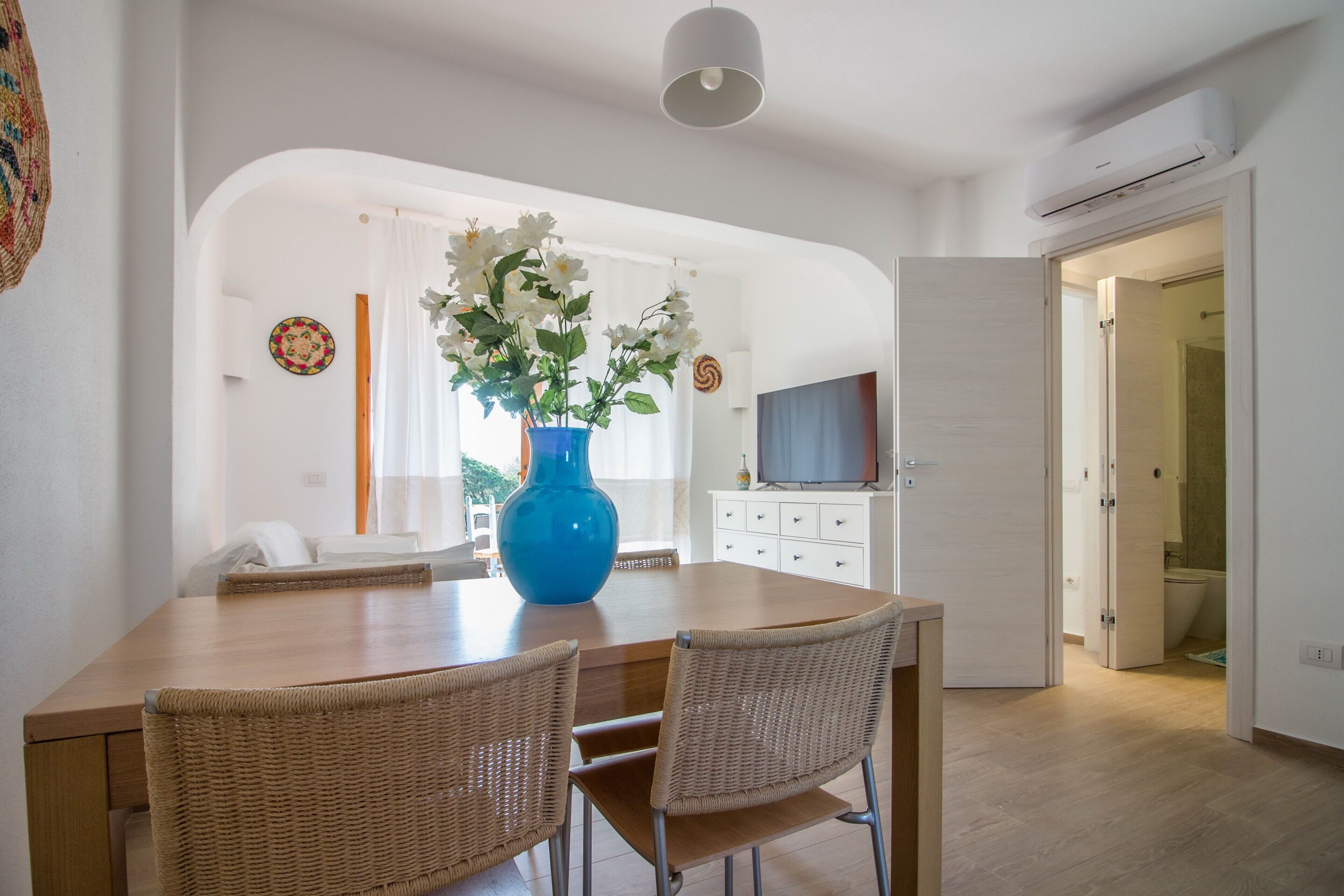 A dining area with a wooden table, wicker chairs, a blue vase with flowers, and a television in the background.