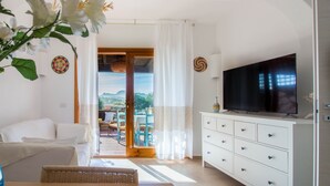 A room with a TV, white furniture, a wicker chair, and a view through a door. - Villa 'Porto Rotondo' with Sea View, Wi-Fi and Air Conditioning (Porto Rotondo)