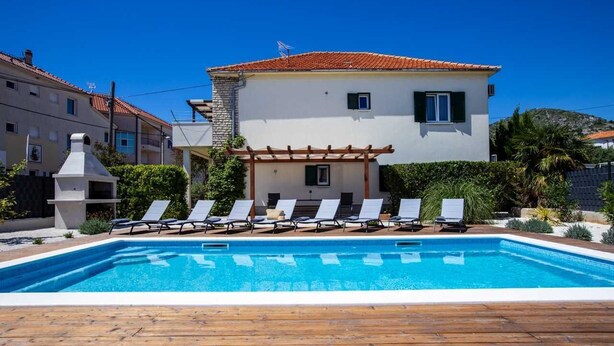Una piscina con sillas de descanso, una pérgola y un edificio blanco con techo rojo.