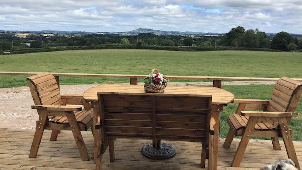Una terraza de madera con una mesa redonda y cuatro sillas, con vistas a un campo cubierto de hierba y colinas lejanas.