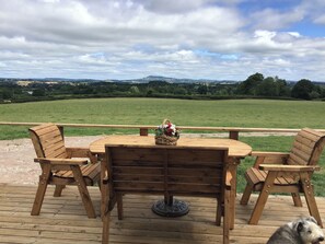 Una terraza de madera con una mesa redonda y cuatro sillas, con vistas a un campo cubierto de hierba y colinas lejanas.