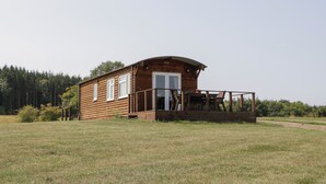 Una cabaña de madera con un porche y una pequeña terraza, rodeada por un campo cubierto de hierba y árboles.