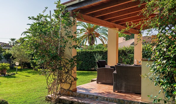 A covered patio area with wicker furniture, a stone pathway, and lush greenery.