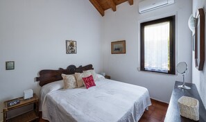 Un dormitorio con cama, una mesita de noche con un reloj, una ventana y un espejo montado en la pared.