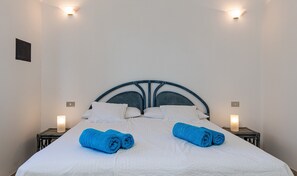 A neatly made bed with white linens and two blue towels, a blue headboard, and wall-mounted lights.