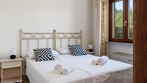 A neatly made bed with white linens and two patterned pillows, a bedside table with a lamp, and a window with curtains. - Villa 'Mariella' with Private Terrace, Wi-Fi and Air Conditioning (Costa Rei)
