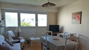A living room with a sofa, a dining table with chairs, a TV, and a window with a view of greenery. - T2 apartment 200 m from the sea (Concarneau)
