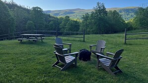 A picnic area with Adirondack chairs and a picnic table, surrounded by a grassy field and forested hills. - Historic Cabin in the Blue Ridge Mountains (Pearisburg)