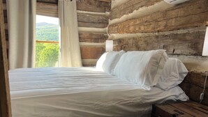 A bed with white linens in a log cabin room with a view of green hills. - Historic Cabin in the Blue Ridge Mountains (Pearisburg)