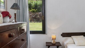 Et soveværelse med et træmøbel, et natbord med lampe, en seng med puder, et vindue med udsigt til en have og en vægmonteret lampe.