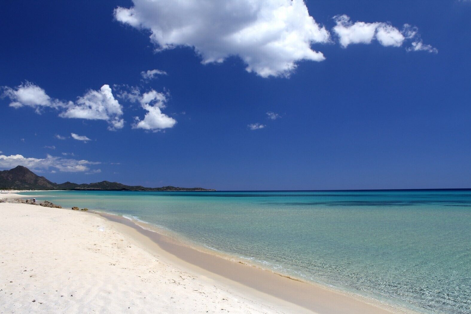 A beach with clear turquoise water, white sand, and distant mountains under a blue sky with clouds.