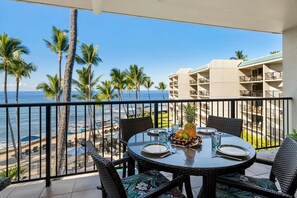 Ein Balkon mit Esstisch, der auf einen Strand mit Palmen und ein Gebäude im Hintergrund blickt.
