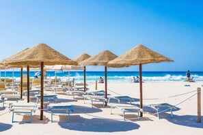 Een strand met ligbedden en rieten parasols, een heldere lucht en de oceaan op de achtergrond.