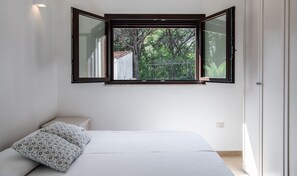 A bedroom with a bed, pillows, a window with a view of trees, and a white wall.