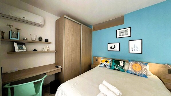 A hotel room with a bed, a desk, a chair, a wardrobe, and wall-mounted shelves.