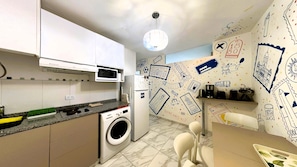 Cozinha compacta com micro-ondas, forno e máquina de lavar. Possui uma área de refeições com mesa e cadeiras. As paredes são decoradas com diversas ilustrações.