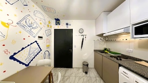 Une petite cuisine équipée d’un four à micro-ondes, d’un évier et d’une poubelle. On y trouve une porte, un calendrier et une horloge fixés au mur.