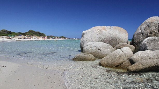 A beach with large rocks, clear water, and a distant shoreline with people. - Villa 'Casa 1' with Sea View, Wi-Fi and Air Conditioning (Castiadas)