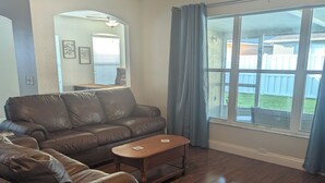 A living room with a brown leather sofa, a wooden coffee table, and a large window with blue curtains. - The Garden House - 3BR /6 Guests (Lutz)