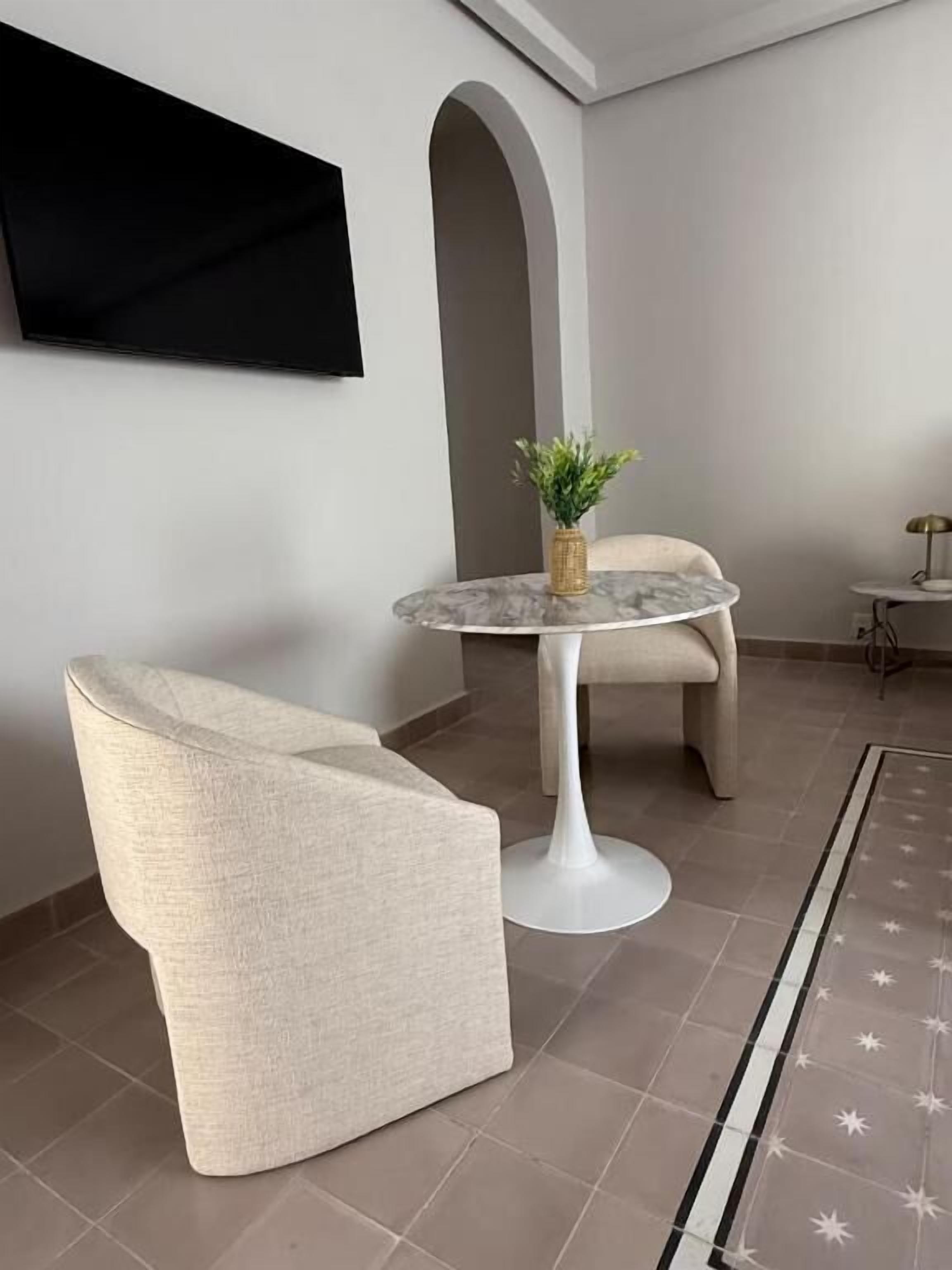 Una sala moderna con una mesa redonda de mármol, un sillón color beige y una mesita de noche con una lámpara.