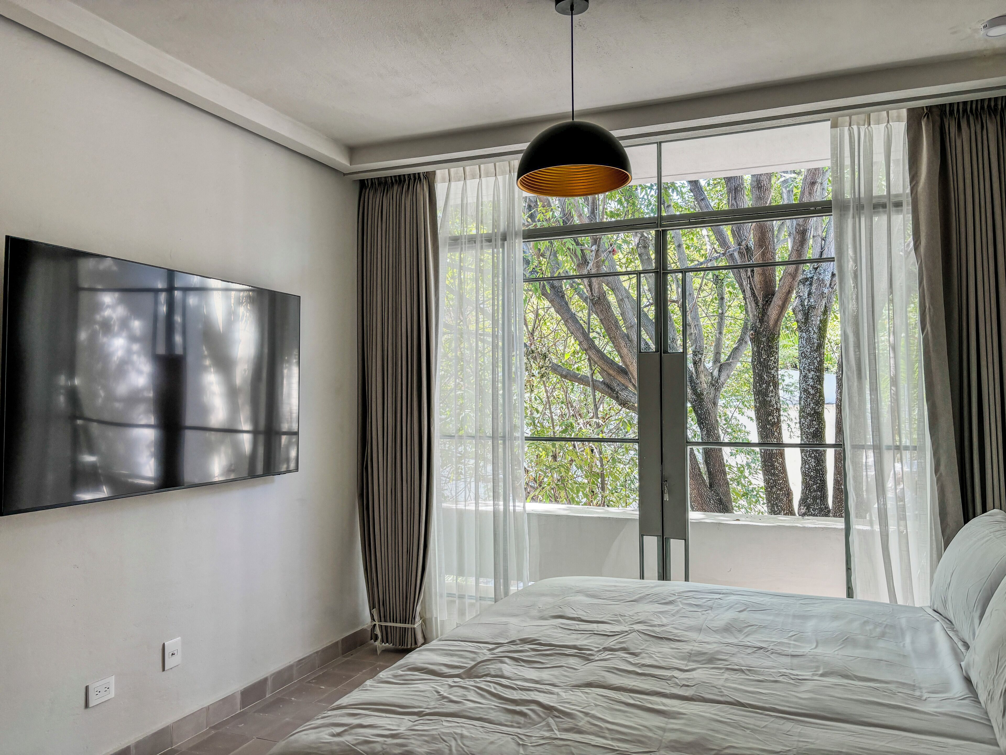 A modern bedroom with a large bed, a flat-screen TV mounted on the wall, and a window with curtains.
