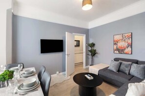 Uma sala de estar moderna com área de jantar, televisão de tela plana na parede, sofá cinza e uma planta em vaso.