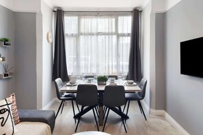 Uma área de refeições com uma mesa posta para quatro pessoas, cadeiras, uma janela com cortinas, uma TV na parede e uma estante com objetos decorativos.
