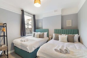 Quarto de hotel com duas camas, janela com cortinas, prateleira com toalhas e uma frase motivacional na parede.