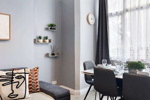 En moderne stue med en sofa, et spiseområde med et bord dækket til fire personer, og et vægur.