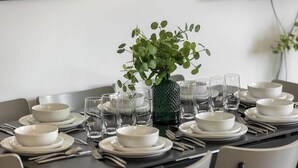 Ett matbord dukat med vita tallrikar, skålar och klara glas, och i mitten står en grön vas fylld med gröna växter.