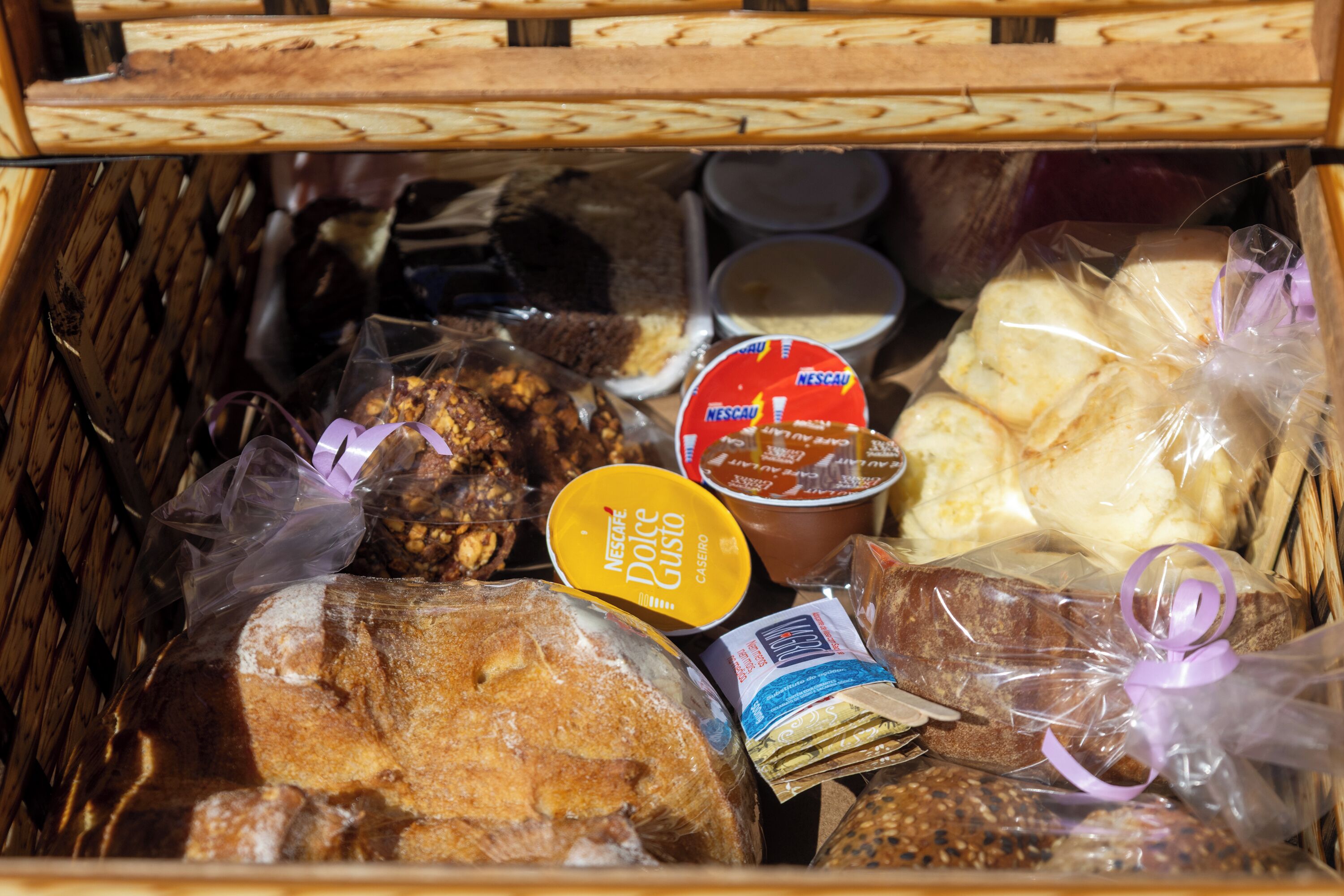 A wicker basket filled with various food items including bread, packaged snacks, and a container of Nescafé Dolce Gusto.