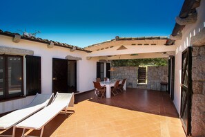 Une terrasse couverte avec une table et des chaises, un coin salon avec des fauteuils et un mur en pierre.