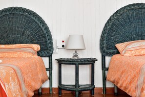 Deux lits en osier avec des têtes de lit assorties, une petite table de chevet avec une lampe et une prise électrique au mur.