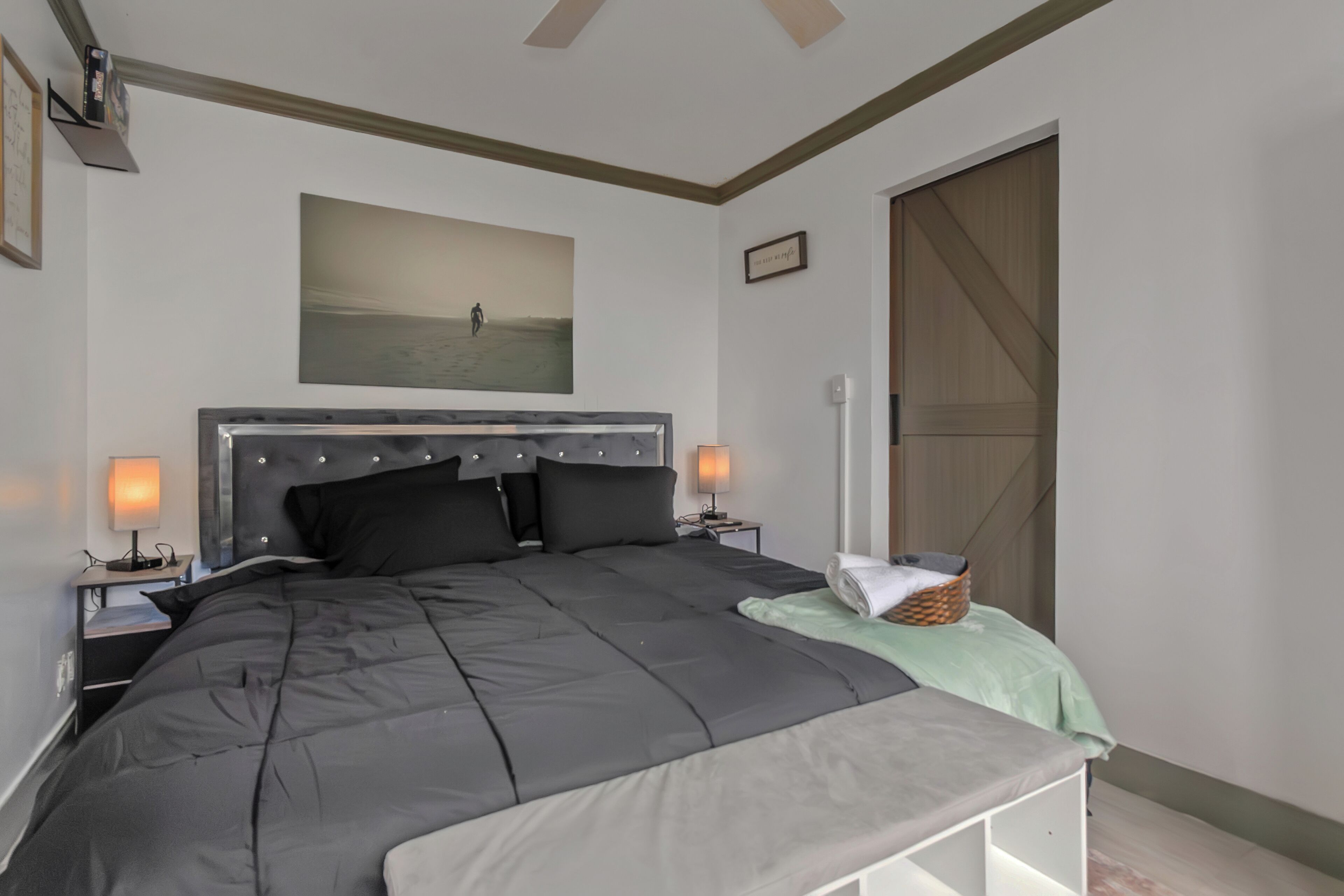 A bedroom with a large bed, two bedside tables with lamps, a headboard, a painting on the wall, a sliding door, and a small basket on the bed.