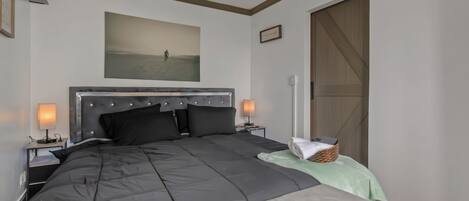 A bedroom with a large bed, two bedside tables with lamps, a headboard, a painting on the wall, a sliding door, and a small basket on the bed.