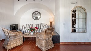 A cozy room with a sofa, two wicker chairs, a coffee table, a TV, a lamp, and a large window with a decorative frame. - Villa 'Casa 2' with Private Terrace, Garden, and Air Conditioning (Castiadas)