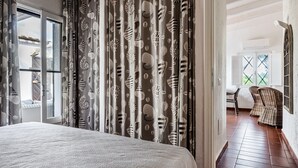 A bedroom with a bed, curtains with a shell pattern, a wicker chair, and a view of the outdoors. - Villa 'Casa 2' with Private Terrace, Garden, and Air Conditioning (Castiadas)