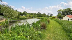 Un sentier paisible le long de la rivière, avec un pont en bois, une végétation luxuriante et un petit bâtiment surmonté d’un toit rouge.
