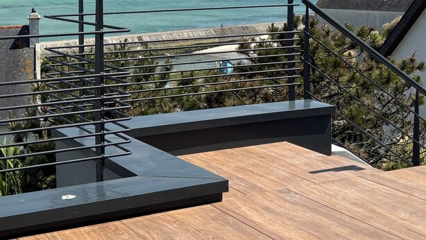 Une terrasse en bois avec une rambarde en métal, donnant sur une zone côtière sous un ciel dégagé.
