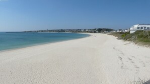 Une vaste plage de sable fin bordée par une mer d’un bleu limpide, quelques bâtiments visibles au loin et un bâtiment blanc avec une enseigne sur le côté droit.