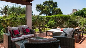 An outdoor patio area with wicker furniture, a glass table, and a view of a garden with trees and shrubs. - Villa 'Salvi' with Private Terrace, Wi-Fi and Air Conditioning (Costa Rei)