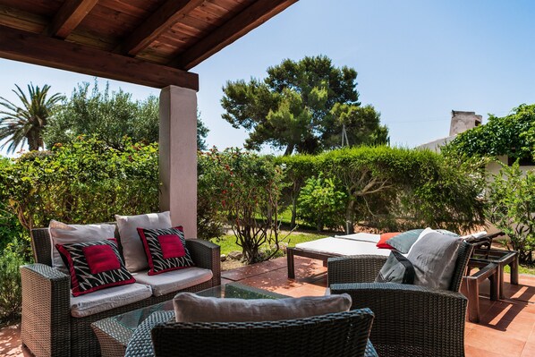 An outdoor patio area with wicker furniture, a glass table, and a view of a garden with trees and shrubs. - Villa 'Salvi' with Private Terrace, Wi-Fi and Air Conditioning (Costa Rei)