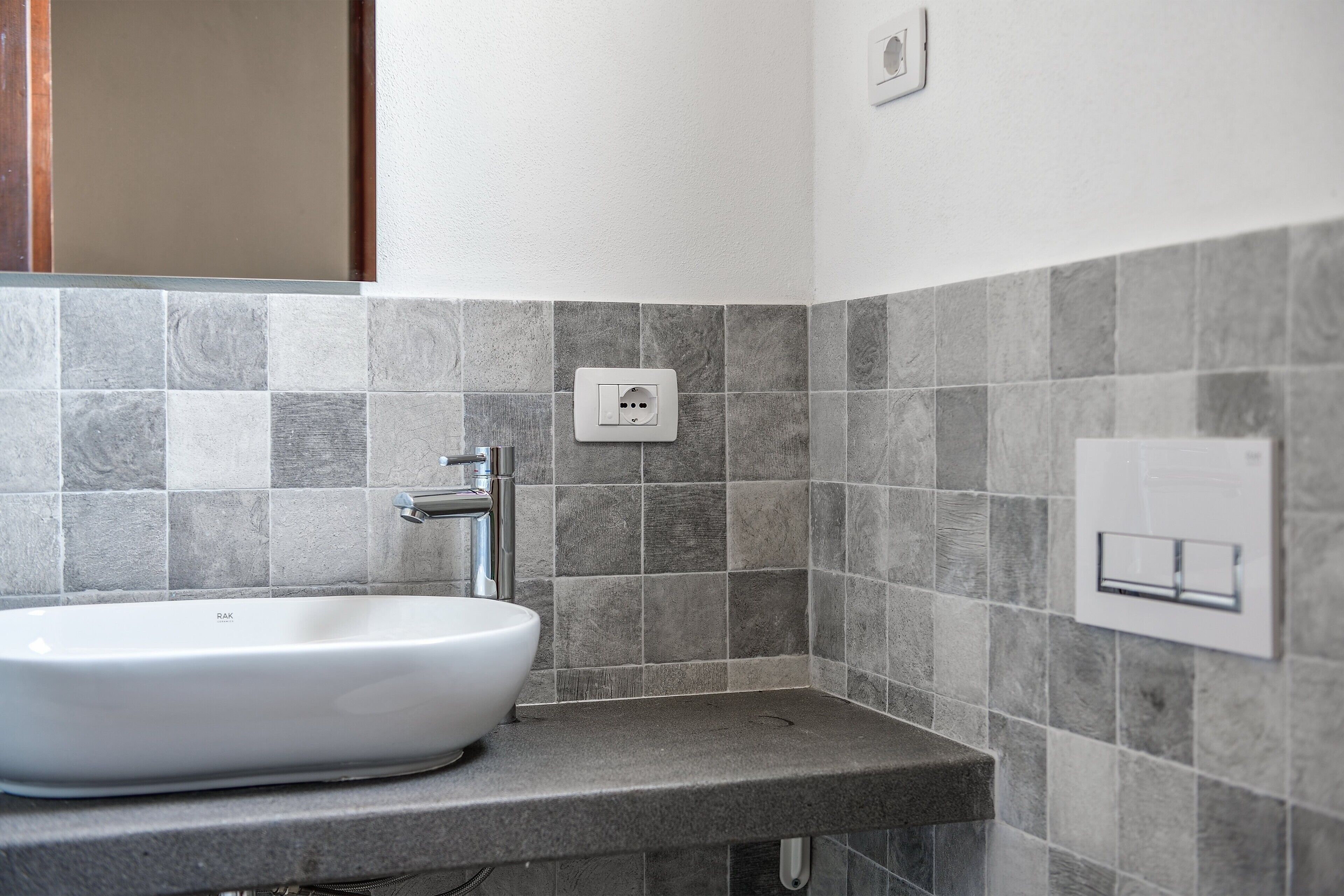 A bathroom with a white sink, grey tiles, a visible electrical outlet, and a wall-mounted light switch.