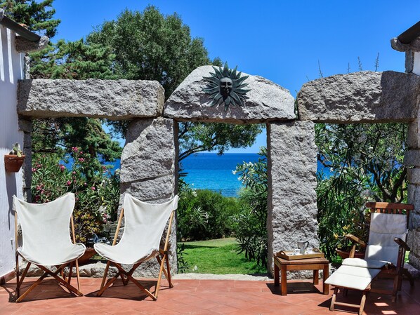 Une arche en pierre ornée d’un motif solaire, deux chaises blanches, une table en bois avec un plateau, et une vue sur la mer.
