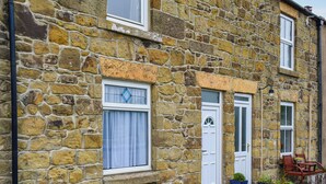 Un bâtiment en pierre avec une porte blanche, une fenêtre avec un rideau et un petit espace extérieur aménagé avec des chaises et des plantes en pot.