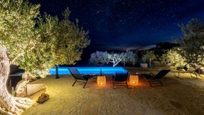 An outdoor pool area with lounge chairs and stone flooring, surrounded by trees and illuminated by night lights. - Holiday Home House of the Sun (Sutivan)