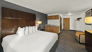 A hotel room with a large bed, a wooden headboard, a desk with a lamp, a kitchenette, and a door leading to another room. - Quality Inn & Suites Caribou (Caribou)