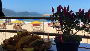 A plate of fried fish with a dipping sauce and lime wedges, a potted plant with red chili peppers, and a view of a beach with umbrellas and people in the background. - Pouso Trindade (Paraty)